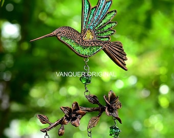 Könnte beinhalten: Ein hängendes Kolibri-Ornament mit einem kupferfarbenen Metallrahmen und schimmernden grünen Glitzereinlagen. Unter dem Vogel hängt ein kupferfarbener Zweig mit drei Blüten, akzentuiert durch baumelnde grüne Glasperlen. Der Text VANQUER ORIGINAL ist sichtbar.