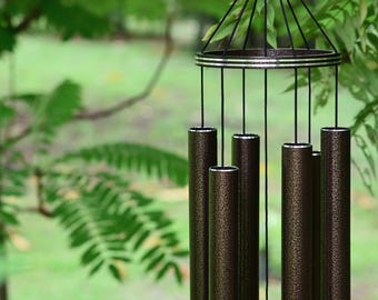 Könnte beinhalten: Ein Windspiel mit sechs zylindrischen Röhren in dunkelbronze-farbener, texturierter Optik. Die Röhren hängen an einem kreisförmigen oberen Rahmen mit silberfarbenen horizontalen Bändern und sind mit schwarzen Schnüren an einem zentralen Aufhängepunkt befestigt.