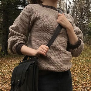 May include: A tan knit sweater with brown trim is paired with a black denim skirt and black tights. A black leather satchel bag is carried over the shoulder. The background features fallen leaves and trees.