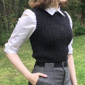 Pode incluir: Um colete de malha preto com textura canelada, usado sobre uma camisa branca de colarinho e calças cinzas com cinto preto.