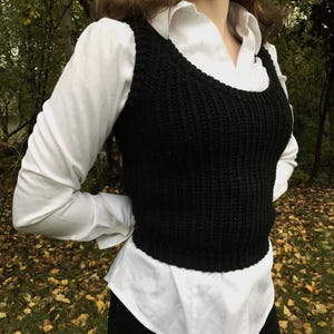 Pode incluir: Um colete preto de malha com decote redondo, usado sobre uma camisa branca com gola. O colete tem uma textura canelada e uma silhueta ajustada. A roupa é completada com calças pretas. O fundo apresenta um ambiente natural ao ar livre.