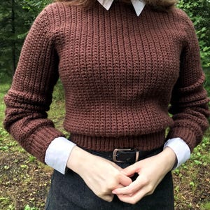 May include: A brown, ribbed-knit sweater with long sleeves. The sweater is worn over a white collared shirt and paired with a dark gray skirt and a black belt with a gold buckle. The sleeves of the shirt are visible.