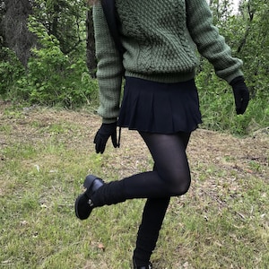 K&ouml;nnte beinhalten: Ein gr&uuml;ner Zopfstrickpullover, ein schwarzer Faltenrock und schwarze Strumpfhosen. Das Outfit umfasst schwarze Handschuhe und schwarze Mary Jane-Schuhe. Die Person tr&auml;gt einen Rucksack und steht auf Gras.