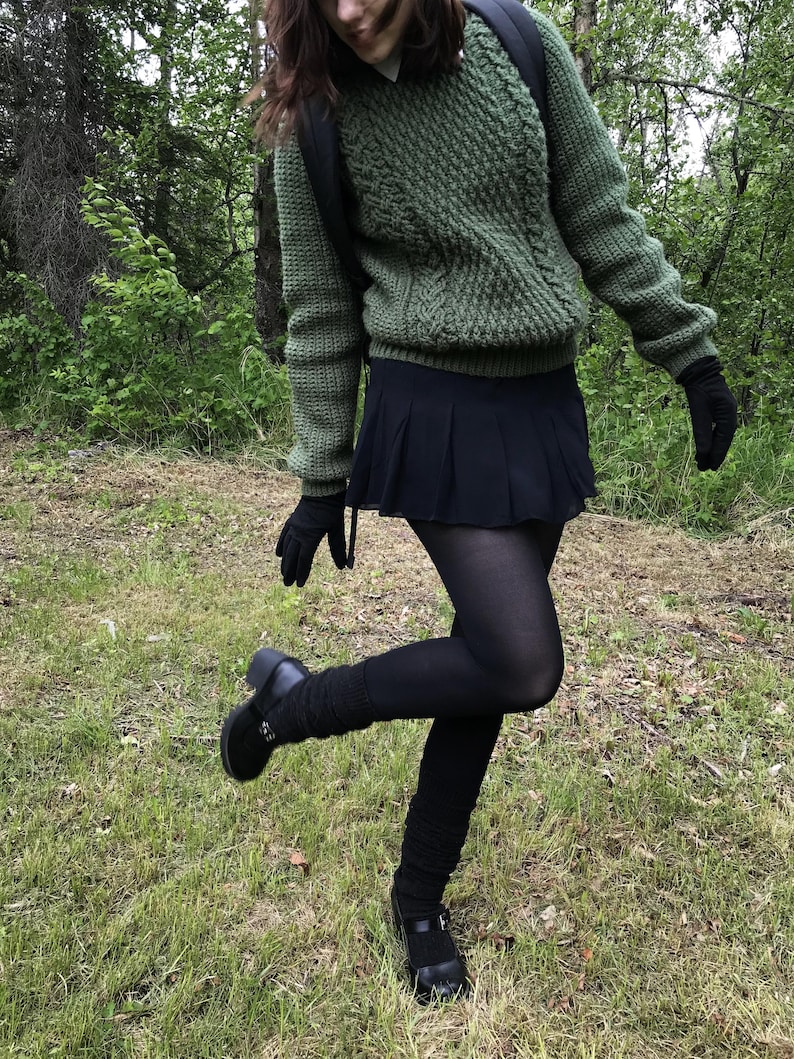 K&ouml;nnte beinhalten: Ein gr&uuml;ner Zopfstrickpullover, ein schwarzer Faltenrock und schwarze Strumpfhosen. Das Outfit umfasst schwarze Handschuhe und schwarze Mary Jane-Schuhe. Die Person tr&auml;gt einen Rucksack und steht auf Gras.