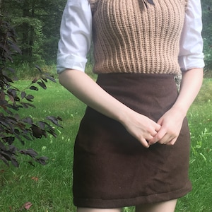 K&ouml;nnte beinhalten: Eine beige, gerippte Strickweste &uuml;ber einem wei&szlig;en Hemd mit Kragen und einer schwarzen Schleife am Hals, kombiniert mit einem einfarbigen, dunkelbraunen Minirock und schwarzen kniehohen Socken. Das Outfit umfasst ein strukturiertes &auml;rmelloses Oberteil.