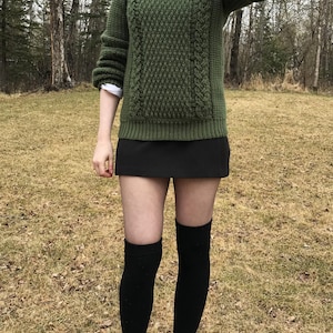 K&ouml;nnte beinhalten: Ein gr&uuml;ner Strickpullover mit strukturiertem Muster und Zopfstrickdetails. Der Pullover wird mit einem schwarzen Rock, schwarzen kniehohen Socken und schwarzen Stiefeln kombiniert. Darunter ist ein wei&szlig;es Hemd mit Kragen zu sehen.