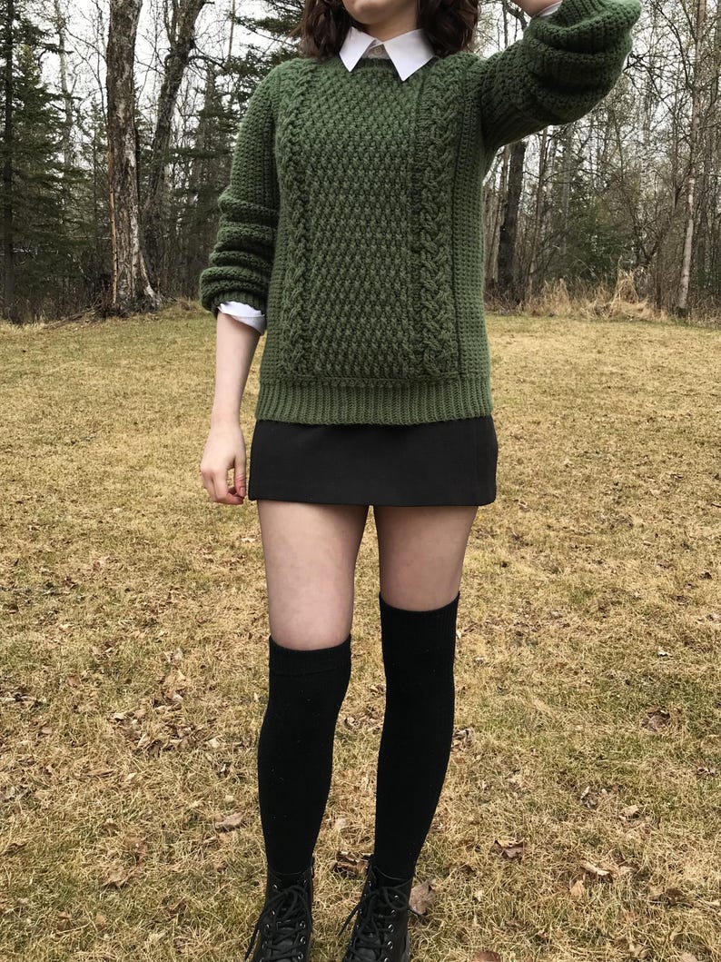 K&ouml;nnte beinhalten: Ein gr&uuml;ner Strickpullover mit strukturiertem Muster und Zopfstrickdetails. Der Pullover wird mit einem schwarzen Rock, schwarzen kniehohen Socken und schwarzen Stiefeln kombiniert. Darunter ist ein wei&szlig;es Hemd mit Kragen zu sehen.