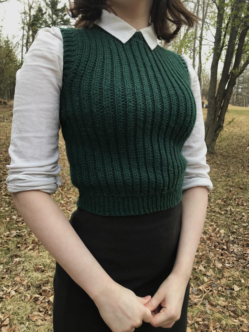 K&ouml;nnte beinhalten: Ein dunkelgr&uuml;ner &auml;rmelloser Strickpullunder mit vertikalem Rippstrickmuster, getragen &uuml;ber einem wei&szlig;en Hemd mit Kragen und langen &Auml;rmeln sowie einem schwarzen Rock. Der Pullunder hat einen Rundhalsausschnitt und eine k&ouml;rpernahe, kurze Passform.