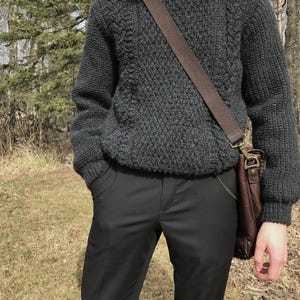 K&ouml;nnte beinhalten: Ein dunkelgrauer Strickpullover mit strukturiertem Muster, kombiniert mit einer schwarzen Hose. Eine braune Ledertasche wird &uuml;ber der Schulter getragen. Ein wei&szlig;es Hemd mit Kragen ist darunter sichtbar.