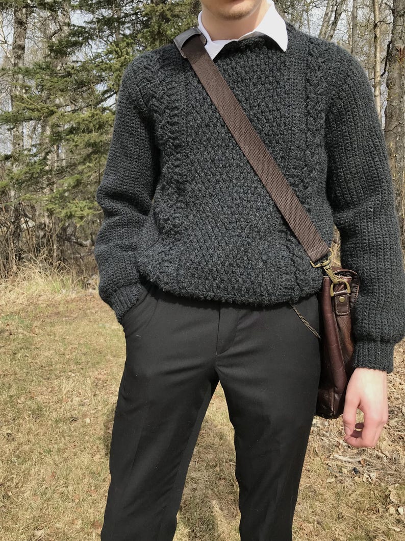 K&ouml;nnte beinhalten: Ein dunkelgrauer Strickpullover mit strukturiertem Muster, kombiniert mit einer schwarzen Hose. Eine braune Ledertasche wird &uuml;ber der Schulter getragen. Ein wei&szlig;es Hemd mit Kragen ist darunter sichtbar.