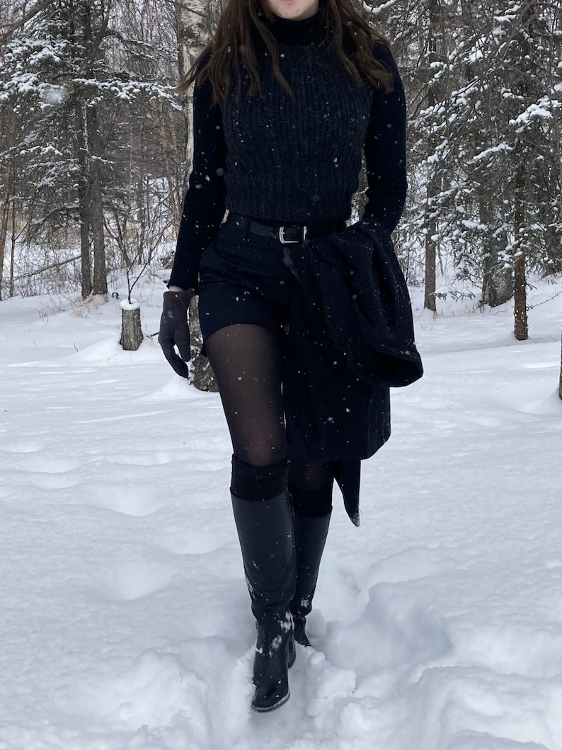 K&ouml;nnte beinhalten: Ein schwarzer Rollkragenpullover, eine dunkel anthrazitfarbene Rippenweste, ein schwarzer G&uuml;rtel, schwarze Shorts, eine transparente schwarze Strumpfhose und kniehohe schwarze Lederstiefel. Ein schwarzer Mantel wird in der Hand gehalten und dunkle Handschuhe.