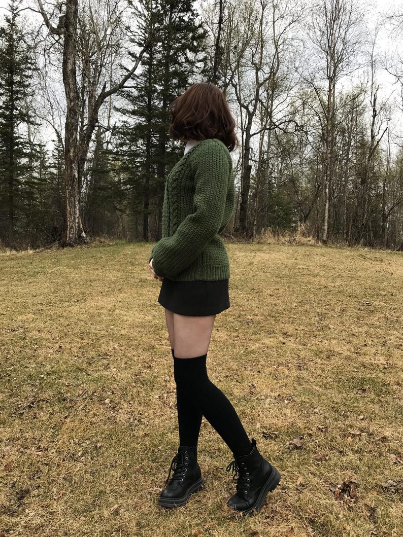K&ouml;nnte beinhalten: Eine Person tr&auml;gt einen gr&uuml;nen Zopfstrickpullover, einen schwarzen Rock und schwarze kniehohe Socken. Die Person tr&auml;gt auch schwarze Kampfstiefel. Der Hintergrund zeigt einen Wald und ein Feld mit trockenem Gras.