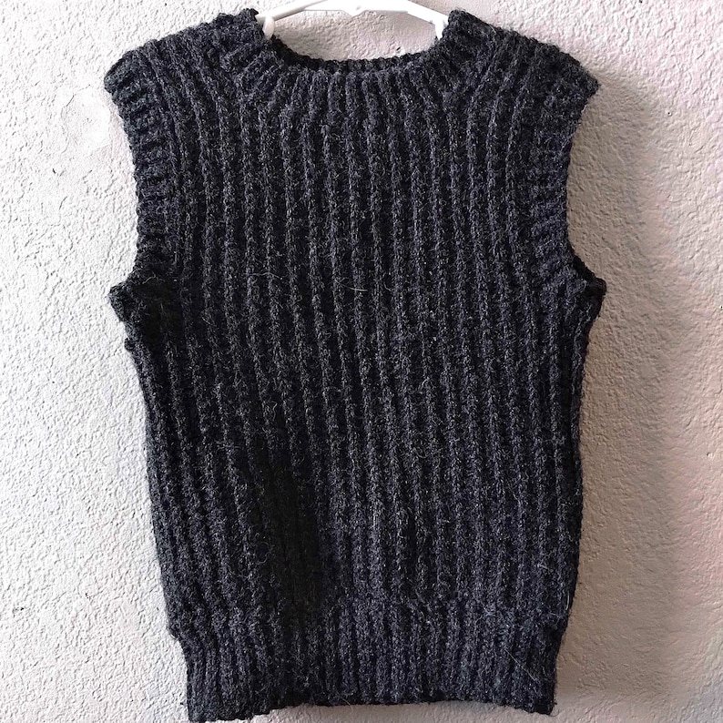 K&ouml;nnte beinhalten: Eine &auml;rmellose Strickweste in Anthrazitschwarz mit einer ausgepr&auml;gten vertikalen Rippenstruktur am gesamten K&ouml;rper. Das Kleidungsst&uuml;ck hat einen Rundhalsausschnitt und einen breiten Rippsaum am unteren Ende aus dickem Garn mit strukturierter Optik.