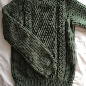 K&ouml;nnte beinhalten: Ein gr&uuml;ner Strickpullover mit Rundhalsausschnitt und langen &Auml;rmeln. Der Pullover hat ein strukturiertes Rautenmuster auf der Vorderseite und Zopfstrickdetails an den Seiten. Die B&uuml;ndchen und der Saum haben ein Rippendesign.