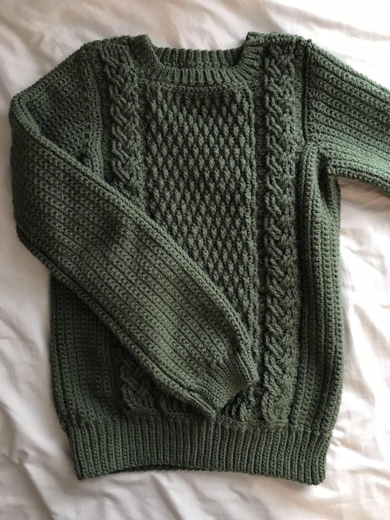 K&ouml;nnte beinhalten: Ein gr&uuml;ner Strickpullover mit Rundhalsausschnitt und langen &Auml;rmeln. Der Pullover hat ein strukturiertes Rautenmuster auf der Vorderseite und Zopfstrickdetails an den Seiten. Die B&uuml;ndchen und der Saum haben ein Rippendesign.