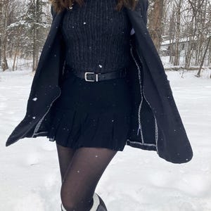 K&ouml;nnte beinhalten: Ein dunkel anthrazitfarbener Rippstrickpullover &uuml;ber einem wei&szlig;en Hemd mit Kragen, kombiniert mit einem schwarzen Faltenrock, einem schwarzen G&uuml;rtel mit silberner Schnalle, blickdichten schwarzen Strumpfhosen und einem langen schwarzen Mantel.