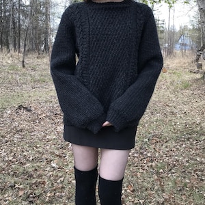 K&ouml;nnte beinhalten: Ein dunkelgrauer, handgestrickter Pullover mit strukturiertem Muster. Der Pullover hat einen Rundhalsausschnitt und lange &Auml;rmel. Die Person tr&auml;gt auch einen schwarzen Rock und schwarze kniehohe Socken. Der Hintergrund ist eine nat&uuml;rliche Umgebung im Freien mit B&auml;umen und Herbstlaub.