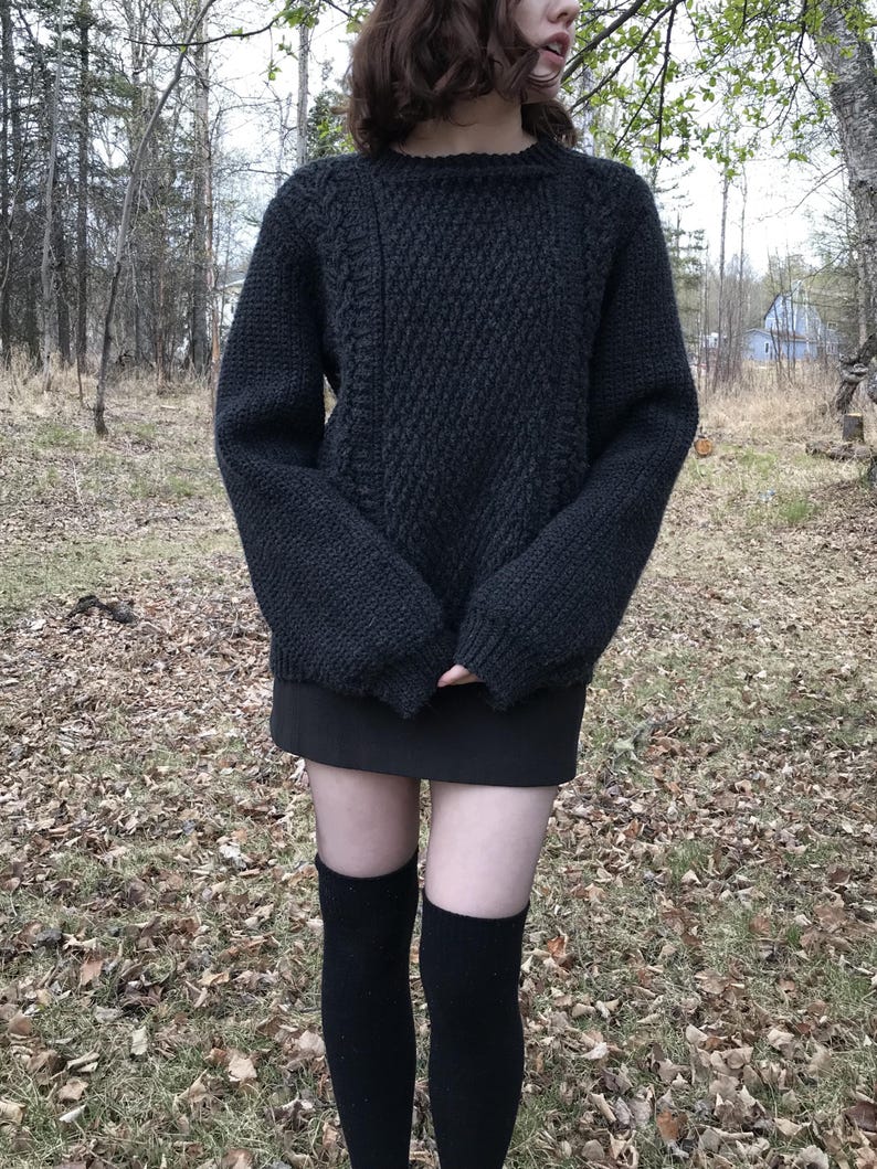 K&ouml;nnte beinhalten: Ein dunkelgrauer, handgestrickter Pullover mit strukturiertem Muster. Der Pullover hat einen Rundhalsausschnitt und lange &Auml;rmel. Die Person tr&auml;gt auch einen schwarzen Rock und schwarze kniehohe Socken. Der Hintergrund ist eine nat&uuml;rliche Umgebung im Freien mit B&auml;umen und Herbstlaub.