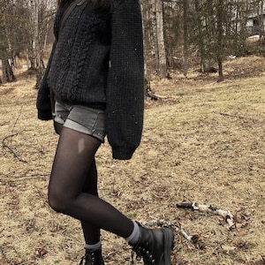 K&ouml;nnte beinhalten: Ein dunkelgrauer Zopfstrickpullover mit hohem Kragen, kombiniert mit grauen Jeansshorts, schwarzen Strumpfhosen und schwarzen Schn&uuml;rstiefeln. Das Outfit wird durch graue Socken und eine braune Umh&auml;ngetasche erg&auml;nzt, vor einem Waldhintergrund.