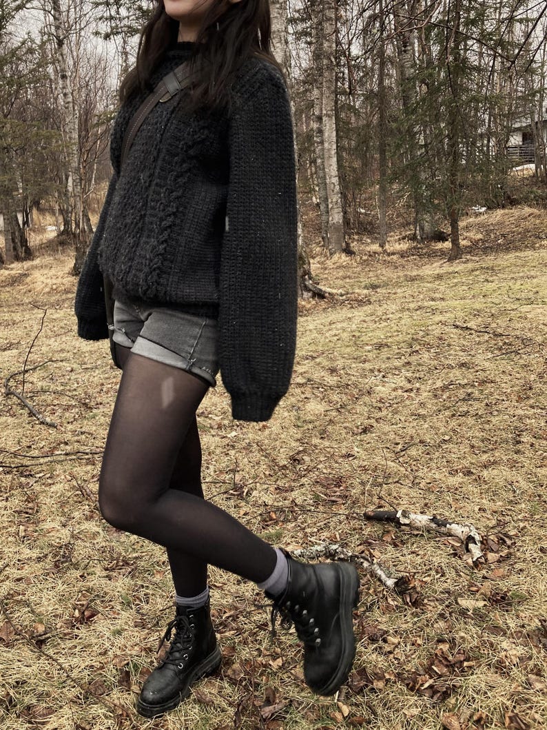 K&ouml;nnte beinhalten: Ein dunkelgrauer Zopfstrickpullover mit hohem Kragen, kombiniert mit grauen Jeansshorts, schwarzen Strumpfhosen und schwarzen Schn&uuml;rstiefeln. Das Outfit wird durch graue Socken und eine braune Umh&auml;ngetasche erg&auml;nzt, vor einem Waldhintergrund.