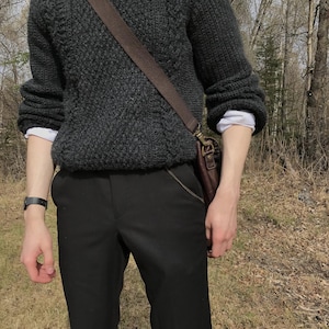 K&ouml;nnte beinhalten: Ein dunkelgrauer, strukturierter Strickpullover &uuml;ber einem wei&szlig;en Hemd mit Kragen. Die Person tr&auml;gt eine schwarze Hose und eine braune Leder-Umh&auml;ngetasche. Eine schwarze Uhr befindet sich am linken Handgelenk.