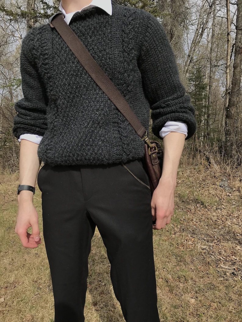K&ouml;nnte beinhalten: Ein dunkelgrauer, strukturierter Strickpullover &uuml;ber einem wei&szlig;en Hemd mit Kragen. Die Person tr&auml;gt eine schwarze Hose und eine braune Leder-Umh&auml;ngetasche. Eine schwarze Uhr befindet sich am linken Handgelenk.