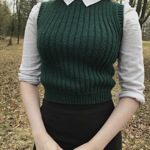 K&ouml;nnte beinhalten: Ein dunkelgr&uuml;ner &auml;rmelloser Strickpullunder mit vertikalem Rippenstrickmuster, getragen &uuml;ber einem wei&szlig;en lang&auml;rmeligen Hemd mit Kragen und einem schwarzen Rock. Der Pullunder hat einen Rundhalsausschnitt und eine k&ouml;rpernahe, kurze Passform.