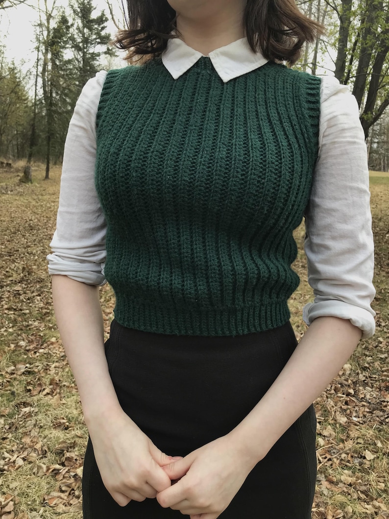 K&ouml;nnte beinhalten: Ein dunkelgr&uuml;ner &auml;rmelloser Strickpullunder mit vertikalem Rippenstrickmuster, getragen &uuml;ber einem wei&szlig;en lang&auml;rmeligen Hemd mit Kragen und einem schwarzen Rock. Der Pullunder hat einen Rundhalsausschnitt und eine k&ouml;rpernahe, kurze Passform.