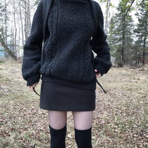 K&ouml;nnte beinhalten: Ein dunkelgrauer, strukturierter Strickpullover mit langen &Auml;rmeln und Rundhalsausschnitt. Kombiniert mit einem schwarzen Minirock und schwarzen kniehohen Socken. Das Outfit wird durch einen schwarzen Rucksack erg&auml;nzt. Der Hintergrund zeigt B&auml;ume und Herbstlaub.
