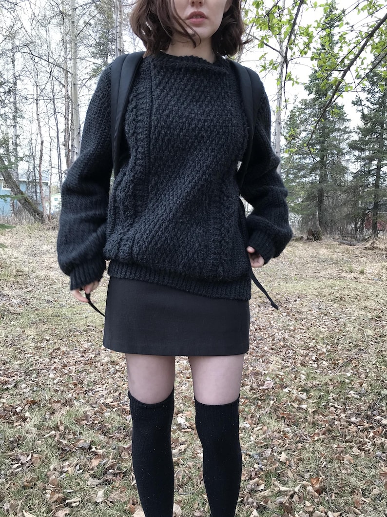K&ouml;nnte beinhalten: Ein dunkelgrauer, strukturierter Strickpullover mit langen &Auml;rmeln und Rundhalsausschnitt. Kombiniert mit einem schwarzen Minirock und schwarzen kniehohen Socken. Das Outfit wird durch einen schwarzen Rucksack erg&auml;nzt. Der Hintergrund zeigt B&auml;ume und Herbstlaub.