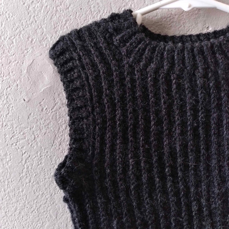 K&ouml;nnte beinhalten: Eine &auml;rmellose Strickweste in Anthrazitschwarz mit einer ausgepr&auml;gten vertikalen Rippenstruktur &uuml;ber den gesamten K&ouml;rper. Das Kleidungsst&uuml;ck verf&uuml;gt &uuml;ber einen dicken, gerippten Rundhalsausschnitt und passende gerippte Armausschnitte aus flauschigem Garn.