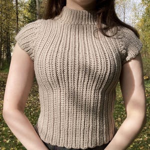 Puede incluir: Un top de ganchillo beige con cuello alto y mangas cortas. La prenda está confeccionada con un patrón de punto acanalado vertical que recorre todo el cuerpo, creando una apariencia texturizada y ajustada en un tono beige neutro.