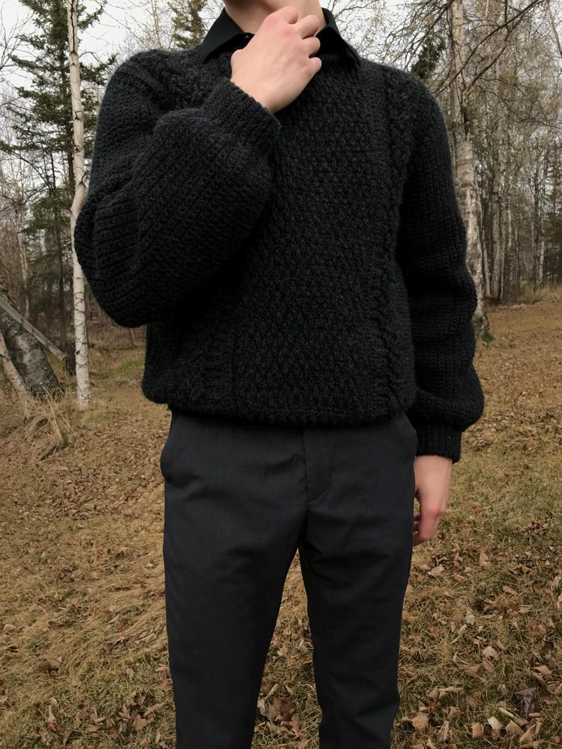 K&ouml;nnte beinhalten: Ein dunkelgrauer, strukturierter Strickpullover mit Rundhalsausschnitt und langen &Auml;rmeln. Der Pullover wird mit einer schwarzen Hose kombiniert. Die Person tr&auml;gt darunter ein schwarzes Hemd mit Kragen. Der Hintergrund zeigt B&auml;ume und Laub.