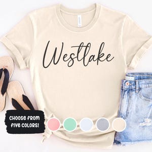 May include: Cream-colored t-shirt with the word "Westlake" in black script. The shirt is paired with denim shorts and sandals. A graphic at the bottom says "CHOOSE FROM FIVE COLORS!" with color swatches.