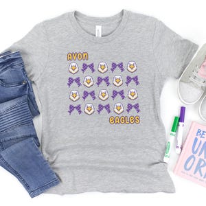 May include: A heather gray t-shirt with the words "AVON EAGLES" in yellow. The shirt features a pattern of purple bows and eagle head graphics. The shirt is displayed with blue jeans, sneakers, and markers.