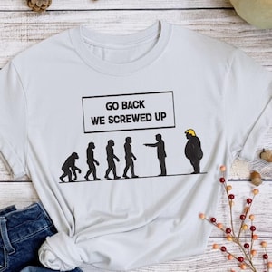 Puede incluir: Una camiseta gris claro con el texto "GO BACK WE SCREWED UP" sobre un gráfico que representa la evolución humana. La última figura de la evolución es una caricatura de una persona con un casco amarillo. Unos vaqueros azules están en primer plano.