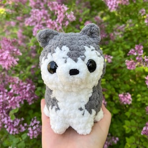 May include: A handmade crochet husky dog plushie. The plushie features a gray and white color scheme, with black eyes and a black nose. The dog is sitting upright, with its ears perked up. The plushie is made of soft, fluffy yarn.