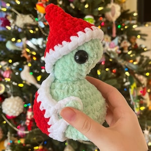 May include: A crocheted green and white toy with a red Santa hat. The toy has a black button eye and is being held in a hand.