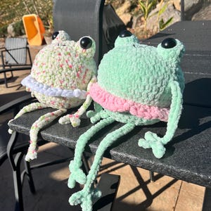 May include: Two crocheted frog plushies sitting on a black table. The frog on the left is white with pink flowers and a white tutu. The frog on the right is green with a pink tutu.
