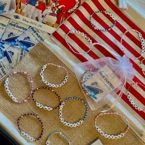 Puede incluir: Una colección de pulseras de cuentas con cuentas de letras blancas que deletrean frases como "Fantasy", "Cruise Era" y "Bon Voyage". Las pulseras están dispuestas con recuerdos de Disney Cruise Line y una tela a rayas rojas y blancas.