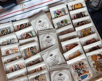 Puede incluir: Una colección de pequeñas bolsas de plástico transparente que contienen varias pegatinas de personajes, dispuestas en filas. Varias bolsas presentan etiquetas blancas circulares con el texto "Hope You Have A Magical Cruise" y "The Herron Family Stateroom 6676".