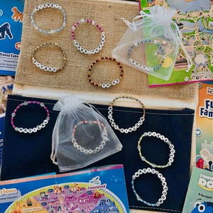Puede incluir: Una colección de pulseras de cuentas con letras blancas que deletrean palabras como "Disney", "Park Hopper" y "Epcot". Las pulseras son de varios colores y se exhiben con mapas de parques de Disney y una pequeña bolsa de regalo.