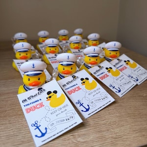 Può includere: Una collezione di paperelle di gomma gialle con cappelli da marinaio bianchi e blu, ciascuna attaccata a un cartellino rettangolare bianco. I cartellini riportano il testo Oh What Luck, You Found A Duck, Keep Or Hide e From Stateroom 6676.