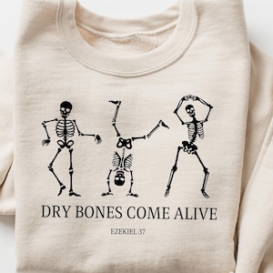 May include: Beige sweatshirt with a black graphic of three dancing skeletons. The text "DRY BONES COME ALIVE" and "EZEKIEL 37" are printed below the skeletons. The sweatshirt has long sleeves and a crew neck.