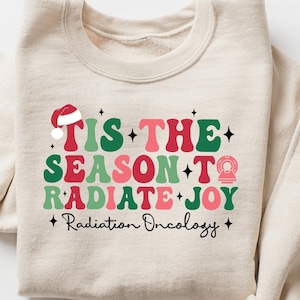Könnte beinhalten: Ein beiger Pullover mit einem weihnachtlichen Motiv, auf dem "Tis the season to radiate joy" mit einem Weihnachtsmann-Hut, einem Weihnachtsbaumschmuck und den Worten "Radiation Oncology" in kleinerer Schrift steht.