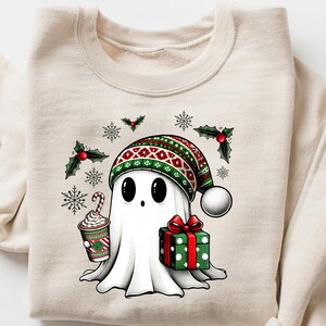 May include: A beige sweatshirt with a white ghost wearing a red and green Christmas hat, holding a cup of hot chocolate and a wrapped gift. The ghost is surrounded by snowflakes and holly.