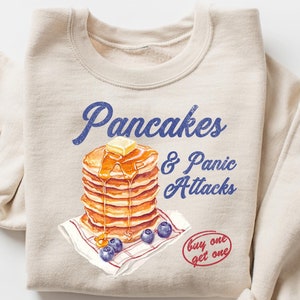 May include: Beige sweatshirt with the text "Pancakes & Panic Attacks" in blue script. The graphic features a stack of pancakes with butter and syrup, blueberries, and a "buy one get one" sticker.