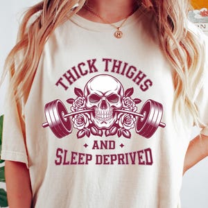 May include: Cream-colored t-shirt featuring a skull graphic with a barbell, roses, and the text "THICK THIGHS AND SLEEP DEPRIVED" in burgundy. The shirt is a casual, comfortable style.