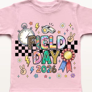 May include: A pale pink t-shirt with the words "FIELD DAY 2026" in vibrant, playful lettering. The design incorporates a stopwatch, whistle, peace symbols, flowers, and lightning bolts, creating a fun, celebratory theme.