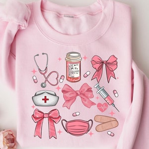 Könnte beinhalten: Rosafarbener Pullover mit einem grafischen Design aus medizinischen Utensilien und rosafarbenen Schleifen. Das Design beinhaltet ein Stethoskop, eine Pillenflasche, eine Spritze, ein Pflaster, eine Gesichtsmaske und eine Krankenschwestermütze.